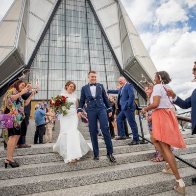 A newlywed couple walks down steps, surrounded by cheering friends and family, outside a modern architectural building under a partly cloudy sky.