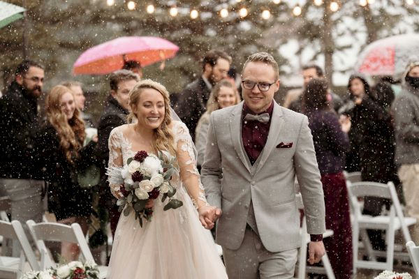 A newlywed couple is walking down an aisle outdoors during snowfall, surrounded by guests, some holding umbrellas.