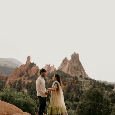 A couple dressed in traditional attire stands hand in hand with a picturesque mountain landscape in the background, surrounded by greenery.