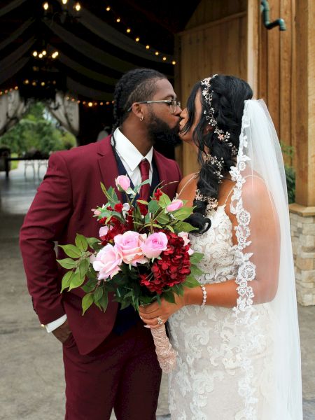 A couple, dressed in wedding attire, share a kiss outdoors. The groom in a maroon suit and the bride in a white gown holding a bouquet.