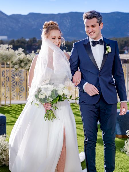 A bride and groom walk outside, with mountains in the background. The bride holds a bouquet and wears a veil.