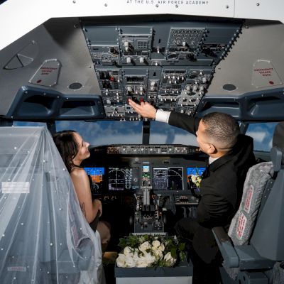 A bride and groom in a flight simulator, with the bride wearing a veil and the groom reaching for controls, surrounded by cockpit panels.