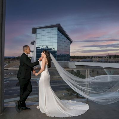 A couple in wedding attire stands on a balcony with the cityscape and a modern building in the background, during a colorful sunset.