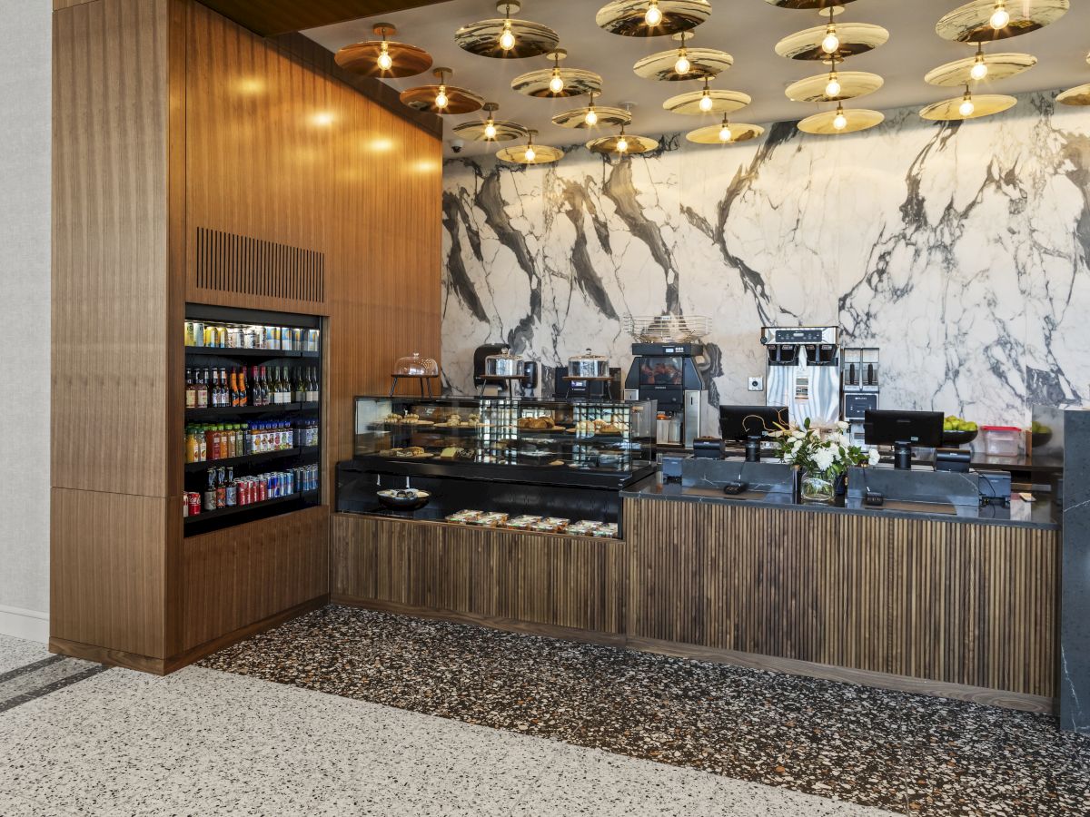 A stylish café corner with a marble backdrop, wooden paneling, self-serve coffee machines, snacks, and decorative light fixtures.