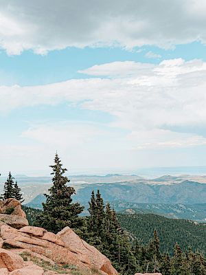 The image shows a scenic mountain landscape with rocky formations, pine trees, and a distant view of rolling hills under a partly cloudy sky.