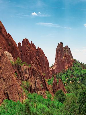 The image shows a landscape of red rock formations with green vegetation and a blue sky with some clouds in the background.