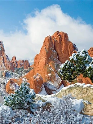 Snow-covered trees and striking red rock formations under a bright blue sky in a scenic landscape.