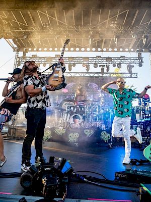 A lively concert with three performers on stage, one raising a guitar, colorful lighting, and music equipment in the background.