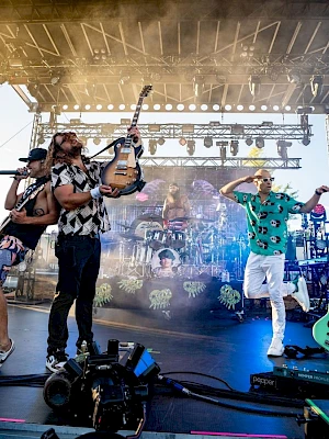 A lively concert with three performers on stage, one raising a guitar, colorful lighting, and music equipment in the background.