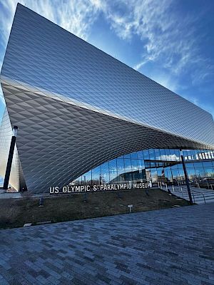 This image shows a modern building with a unique geometric design. The entrance is identified as the U.S. Olympic & Paralympic Museum.