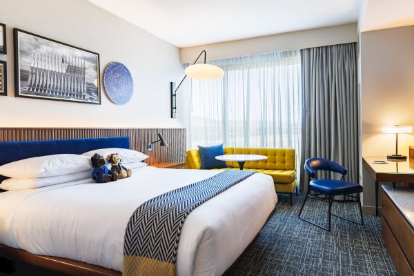 Modern hotel room with a large bed, yellow sofa, blue chair, desk, and window with curtains. Artwork decorates the walls.