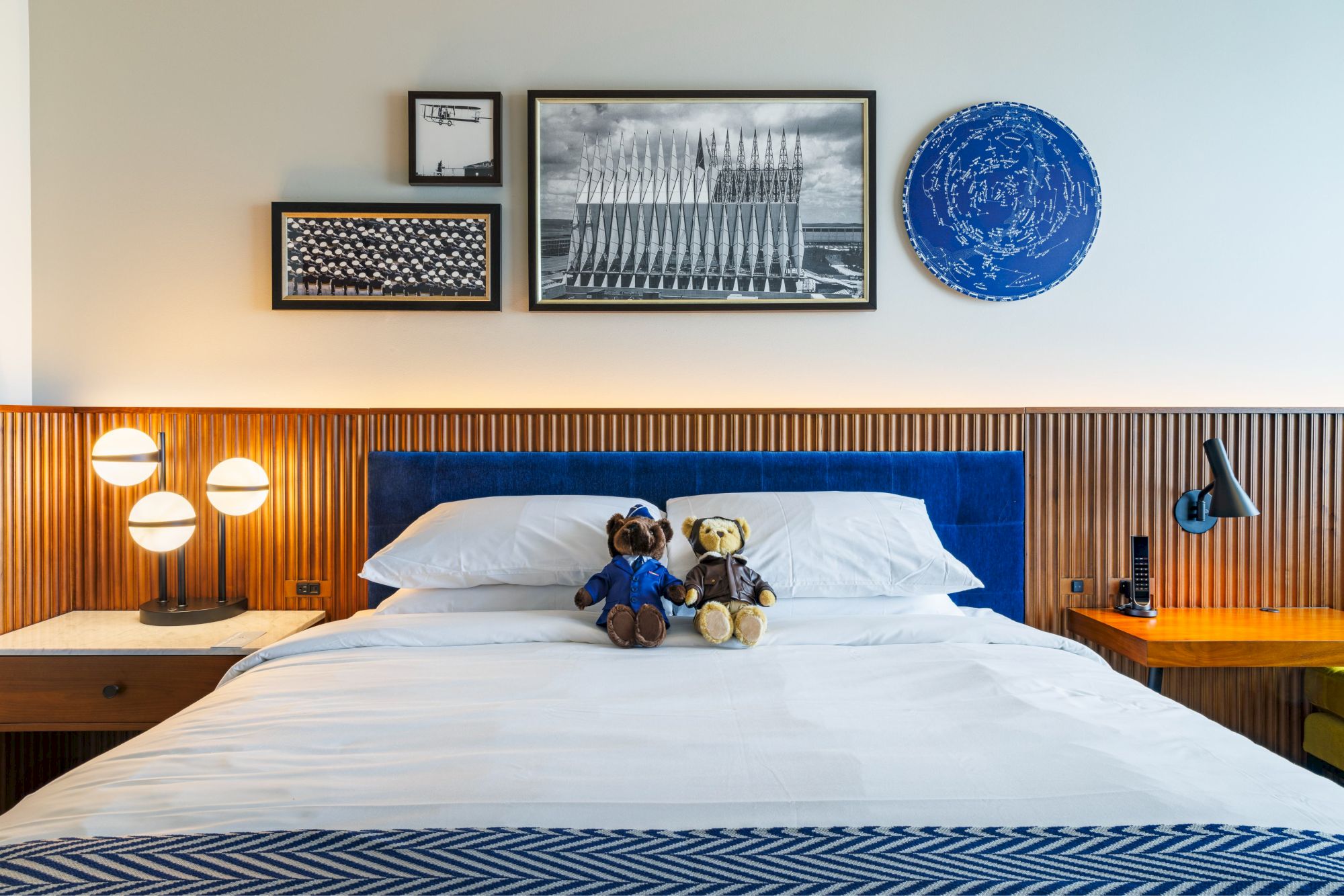 A neatly made bed with two teddy bears, flanked by nightstands, and art above. Unique lamps add a touch of modern style to the room.