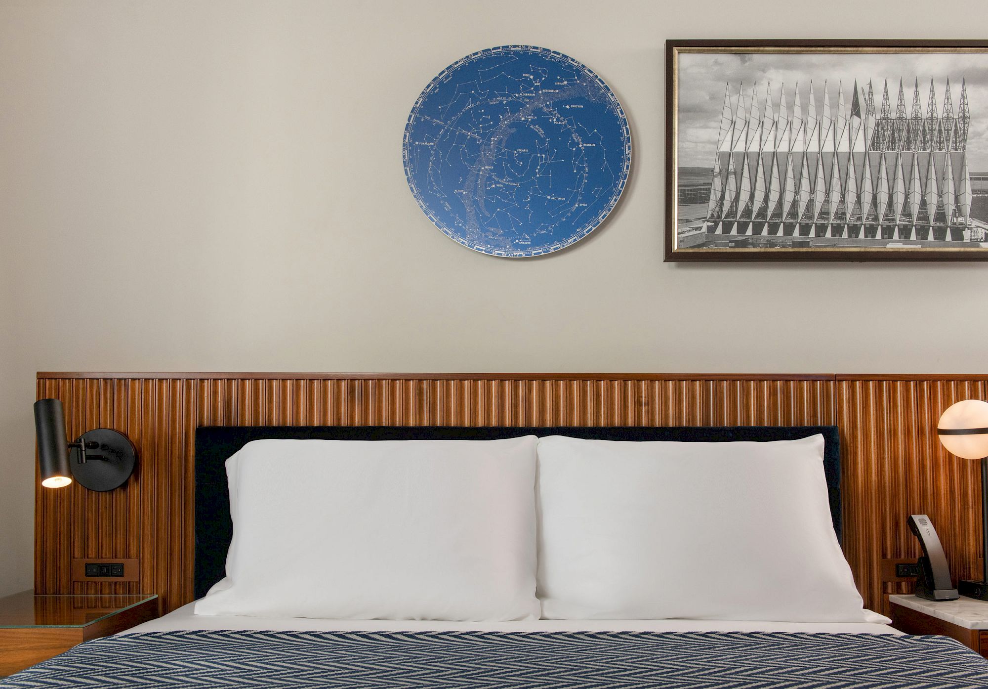 A neatly made bed with white pillows, flanked by black wall lamps, and two framed artworks above: a blue star map and a monochrome architectural image.