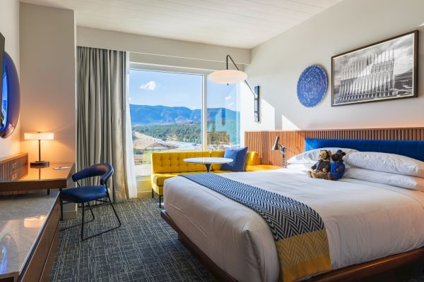 A cozy hotel room with a large bed, desk, yellow sofa, and window view of mountains. Simple modern decor enhances the ambiance.