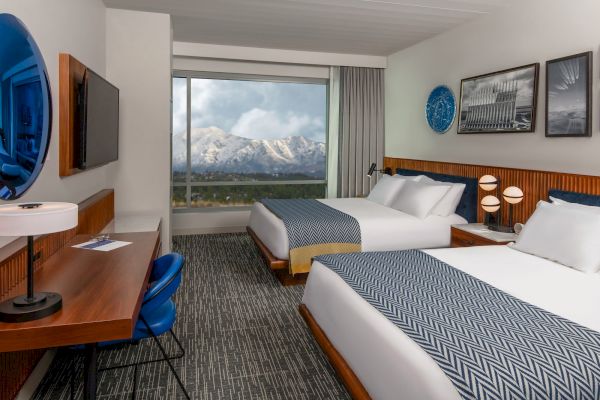 A hotel room with two beds, a desk, chair, TV, and wall art. A large window shows a view of snow-capped mountains.