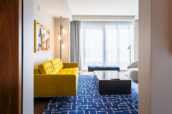 A modern living room features a yellow sofa, geometric rug, and large windows with sheer curtains, creating a bright atmosphere.