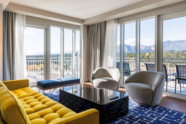 A modern living room with a yellow couch, two gray chairs, a black coffee table, and large windows showing a mountain view.