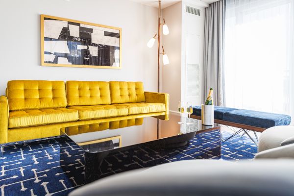 A modern living room with a yellow sofa, abstract art, a blue bench, and a geometric rug. A table holds a champagne bottle and glass.