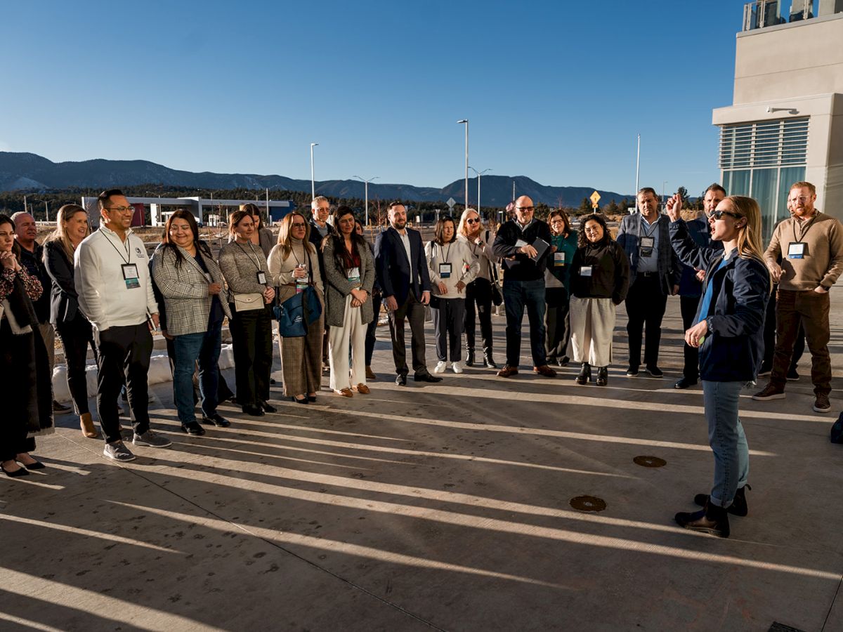 A group of people stands outdoors, listening to a speaker, with mountains in the background under a clear blue sky.