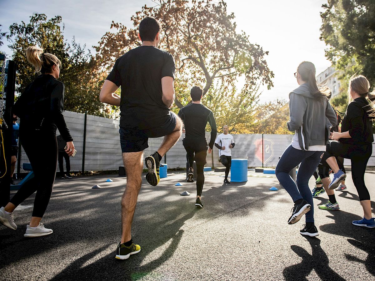 A group of people exercising outdoors in a sunny park, performing leg lifts, wearing athletic clothing, surrounded by trees and buildings.