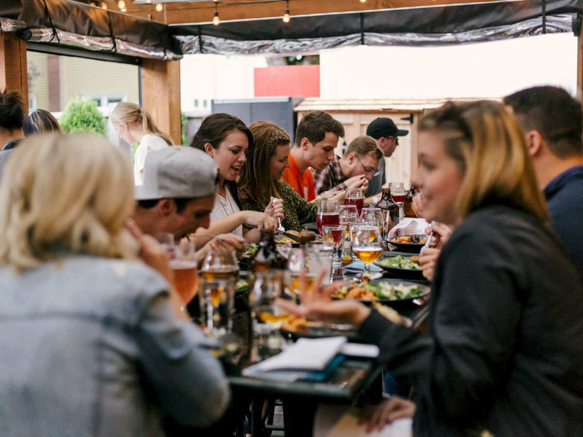 A group of people is enjoying a meal together at an outdoor dining area, engaging in conversation and laughter, with food and drinks on the table.