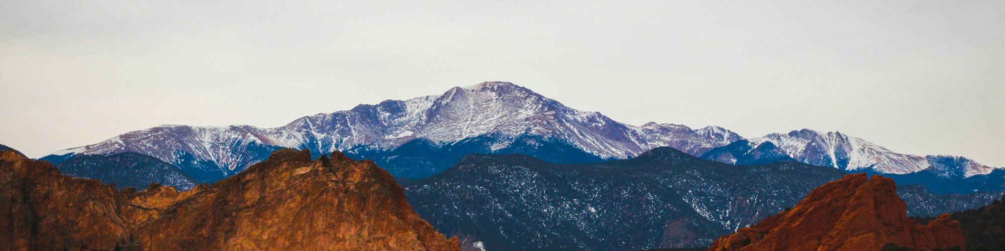 A scenic mountain range with snow-capped peaks, surrounded by red rocks and green vegetation on the hills in the foreground.