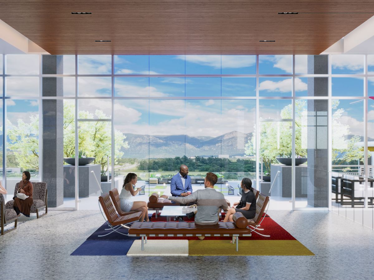 People are seated in a modern lounge area with large windows, mountains and a blue sky in the background, having a discussion.