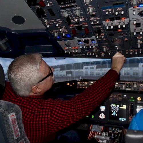 A person wearing a red shirt is operating controls in an airplane cockpit, adjusting switches on the overhead panel.