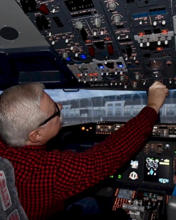 A person wearing a red shirt is operating controls in an airplane cockpit, adjusting switches on the overhead panel.