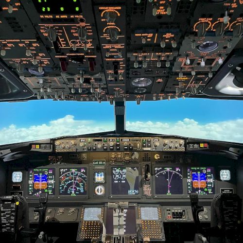 This image shows the cockpit of an aircraft, with various control panels, switches, and displays, and a view of the blue sky and clouds outside.