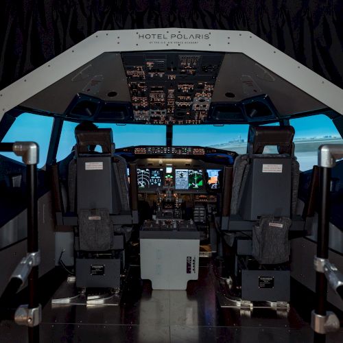 The image shows an airplane cockpit with two pilot seats, control panels, and simulated flight displays, under the label 