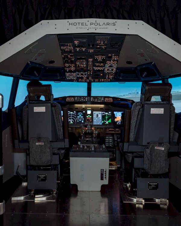 The image shows an airplane cockpit with two pilot seats, control panels, and simulated flight displays, under the label 