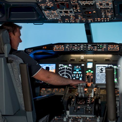 A pilot operating an aircraft cockpit, surrounded by various controls, screens, and instruments, indicating active flight operations.