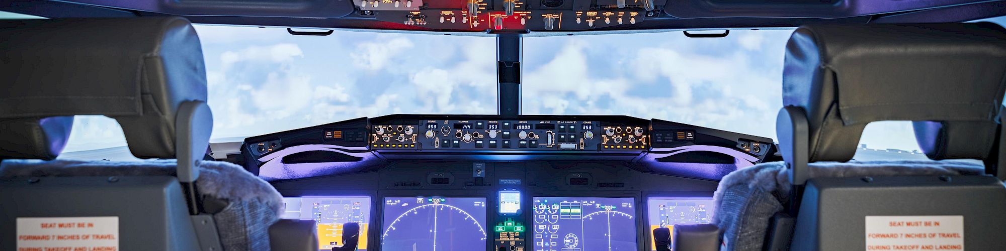 The image shows an aircraft cockpit with control panels and displays, featuring seats for pilots against a backdrop of a cloudy sky.