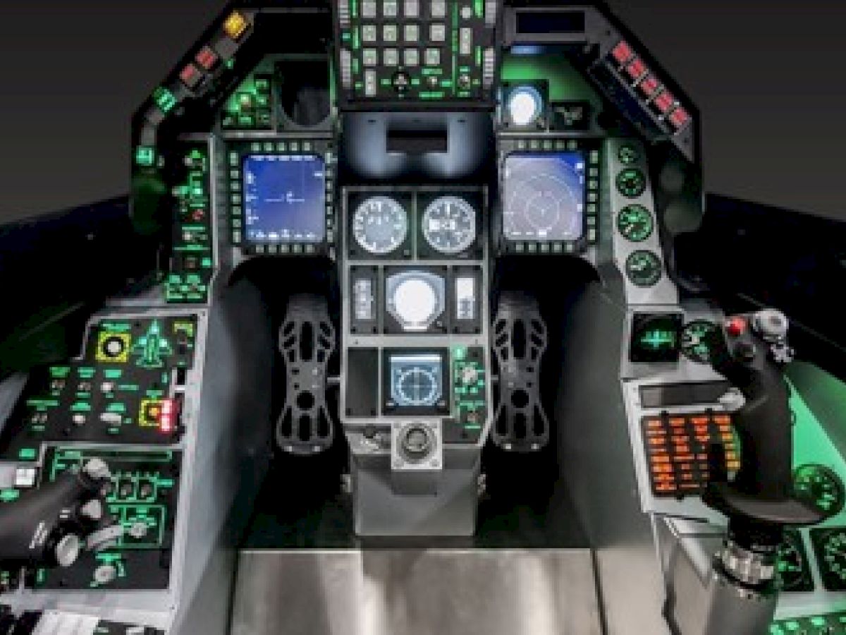 The image shows the cockpit of a modern military aircraft, featuring multiple digital displays, various control switches, and joystick controls.