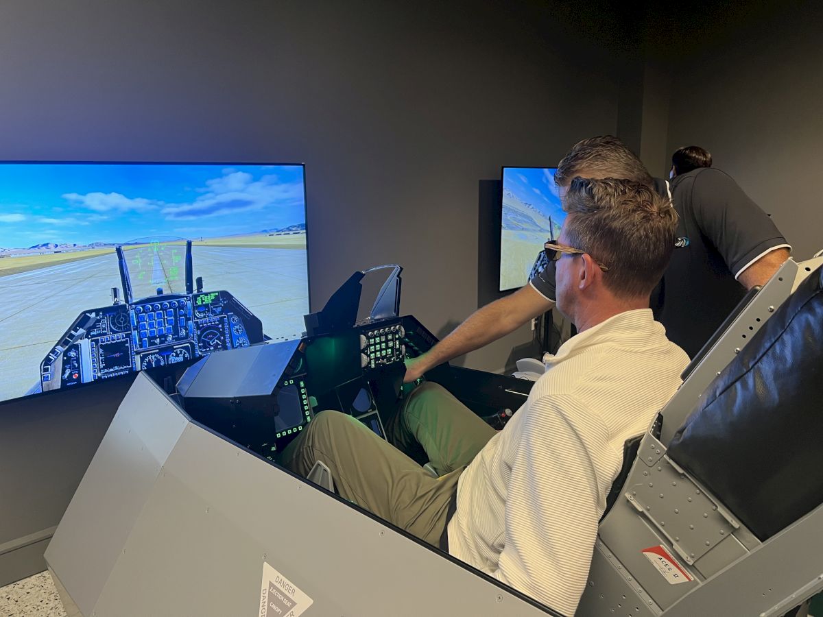 Two people are interacting with a flight simulator setup, featuring a cockpit-style seat and large screen displaying aircraft controls.