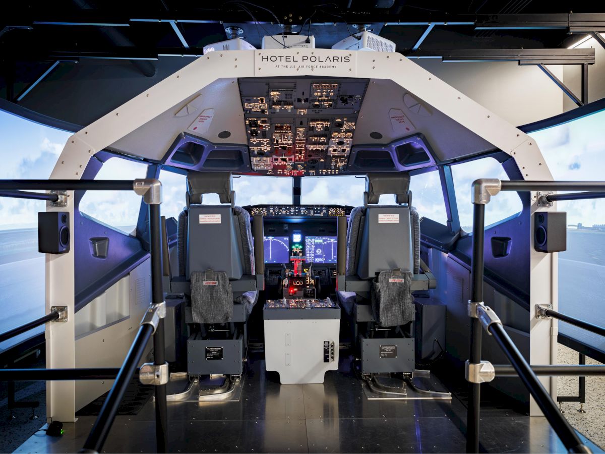 This image shows the interior of an aircraft flight simulator cockpit, complete with seats, controls, and a screen displaying sky and runway.