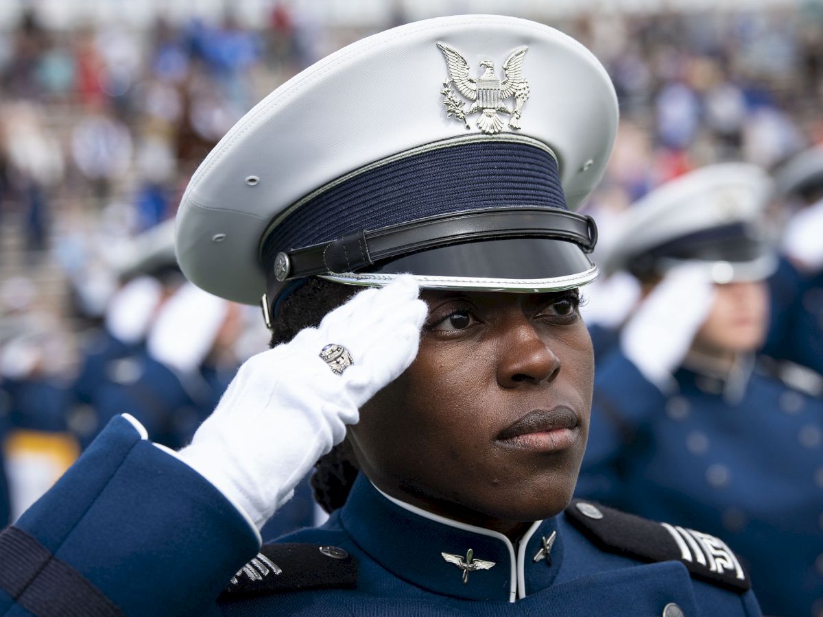 A person in a military uniform is saluting, wearing a white hat and gloves, with others in similar attire blurred in the background.