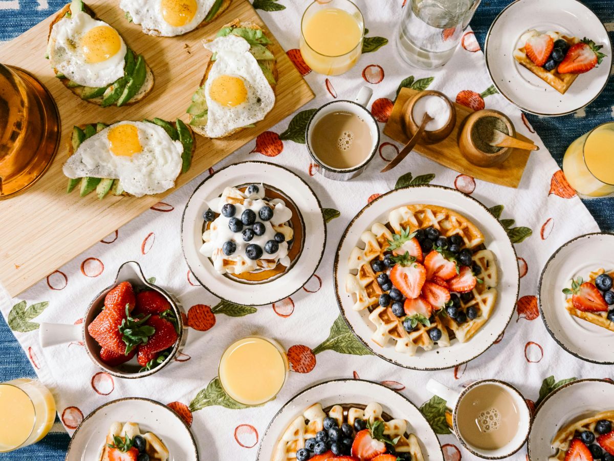 A breakfast spread with waffles topped with berries, eggs on toast, fresh strawberries, and glasses of orange juice on a patterned tablecloth.