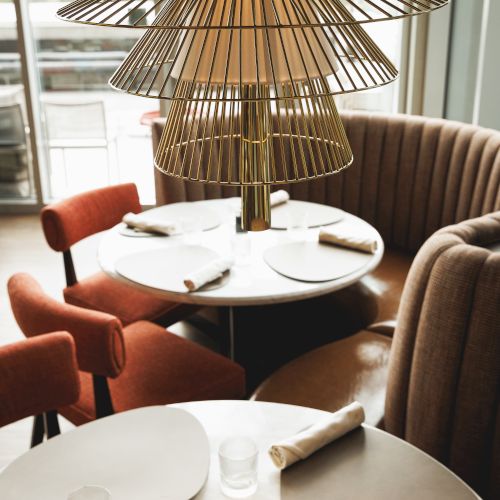 A cozy restaurant interior with round tables, red cushioned chairs, and elegant lighting, set for dining with placemats and glasses.