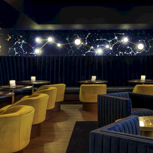 The image shows a stylish, dimly lit lounge with yellow chairs and dark sofas, featuring constellation-themed wall décor.