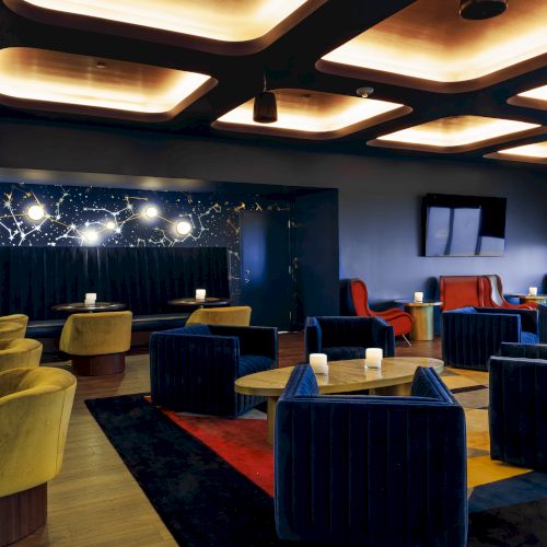 A modern lounge with colored chairs, round tables, and dim lighting. The ceiling has unique panels, and there's a large screen on the wall.