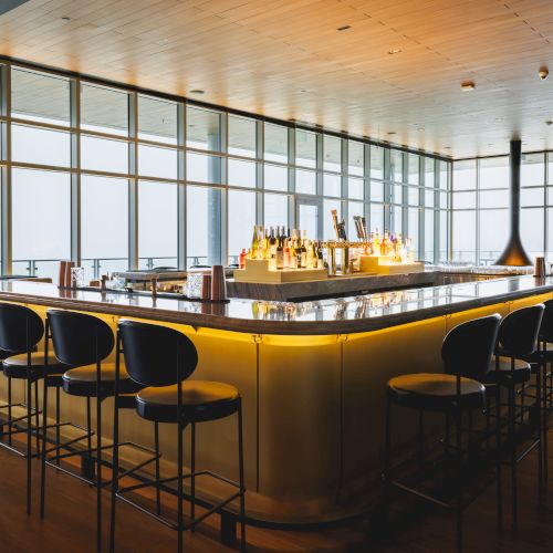 A modern bar with a sleek counter, lined with stools and illuminated bottles, set in a room with large windows and wooden flooring.