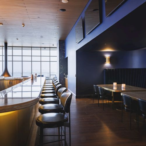 A modern bar interior with a sleek wooden counter, bar stools, dim lighting, and a cozy booth seating area with candles on the tables.