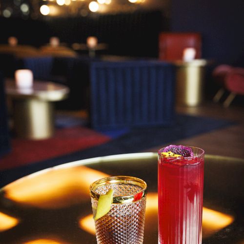 The image shows two cocktails on a table in a dimly lit bar with elegant decor and soft lighting.