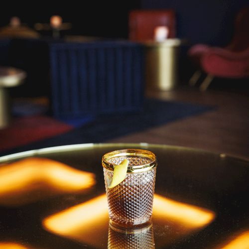 A cocktail with a lemon twist sits on a reflective table in a dimly lit lounge with blurred background furnishings and warm lighting.