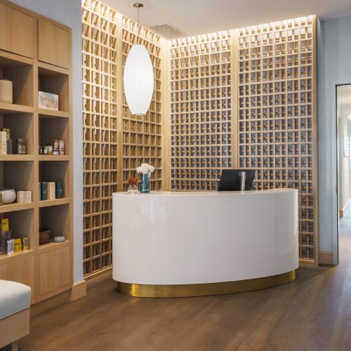 The image shows a modern reception area with a white curved desk, wooden wall accents, shelving with products, and a hallway in the background.