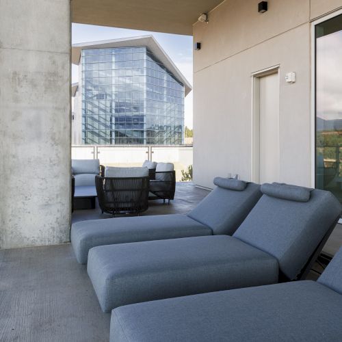 A modern rooftop patio with lounge chairs and a glass-walled building in the background, under a partly cloudy sky.