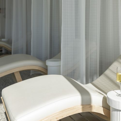 A relaxing spa scene with white chaise lounges, sheer curtains, and a glass of champagne on a small table, creating a serene atmosphere.