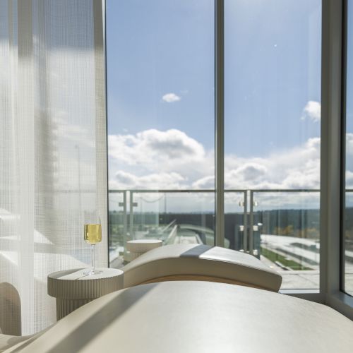 A relaxing lounge area with a curved chair, a side table with a glass, and large windows offering a view of a sunny sky and landscape.